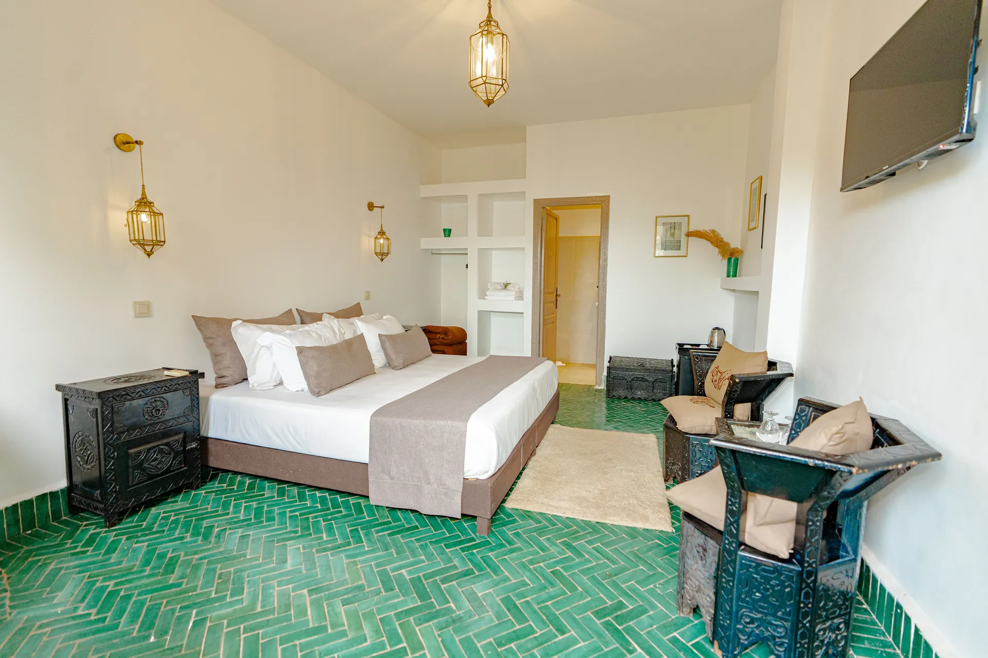 Chambre standard aux murs blancs, lit double drapé de blanc et de coussins beiges, coffres berbères en bois peint et sol en zellige vert