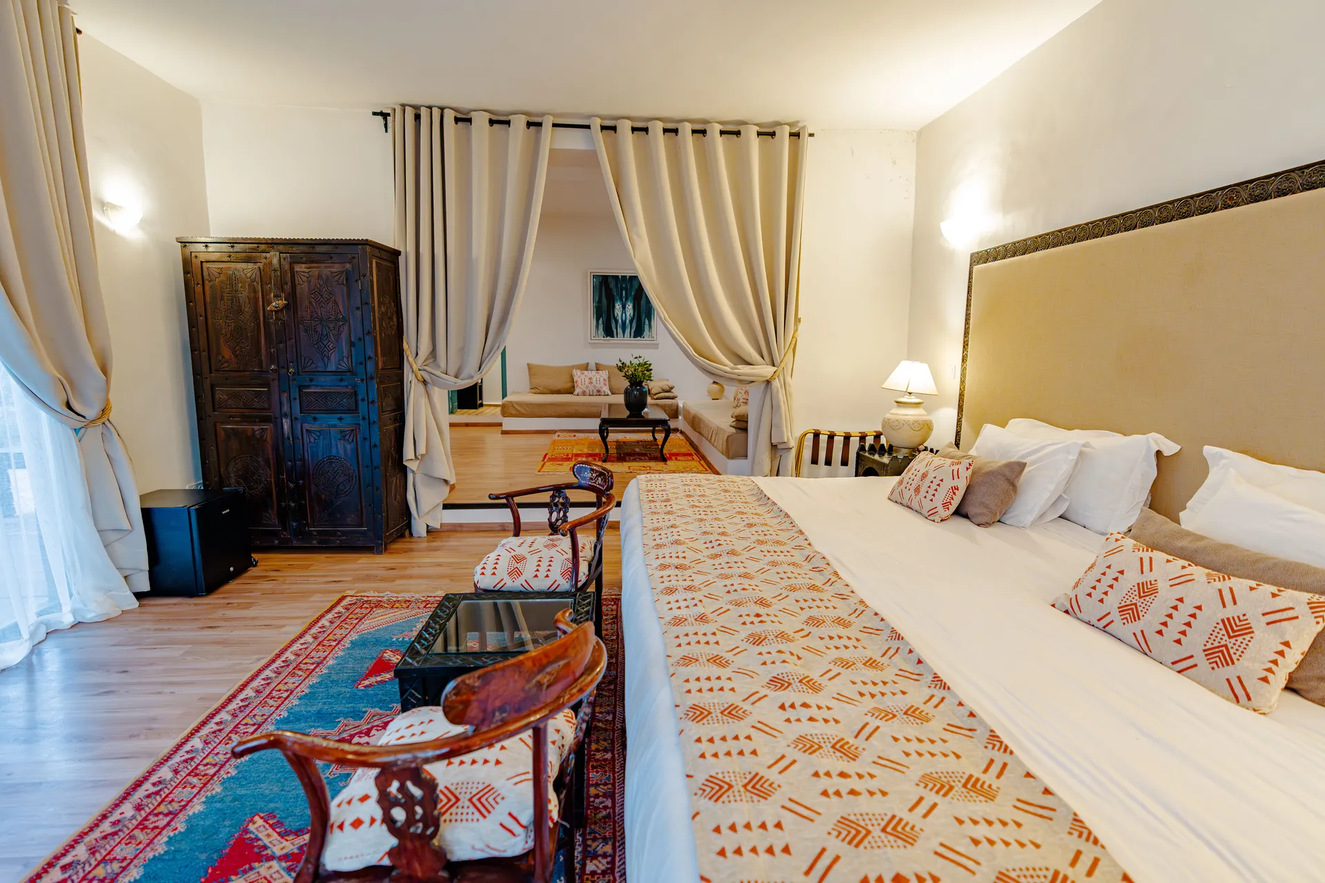 Suite Kasbah avec lit habillé d'un jeté brodé, armoire ancienne en bois sculpté et salon en enfilade derrière des rideaux drapés