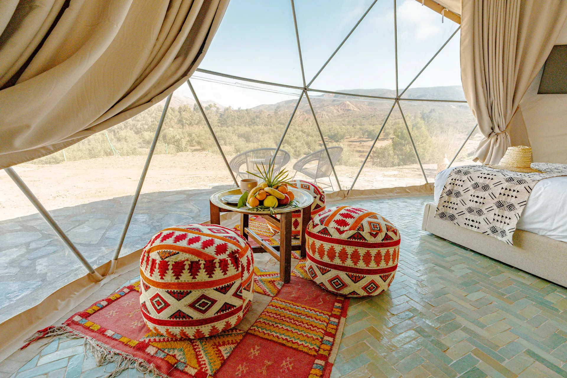Interieur d'un dome Atlas, poufs berberes brodes et tapis rouge sur un sol en zellige, baie geodesique ouverte sur les collines et les oliviers